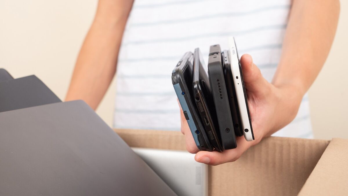 Dłoń trzyma kilka starszych modeli telefonów komórkowych nad otwartym kartonem. Urządzenia są ułożone jedno na drugim i różnią się kolorem oraz grubością obudowy. W kadrze widoczne są także kartonowe pudełko oraz fragment sylwetki osoby trzymającej telefony.
