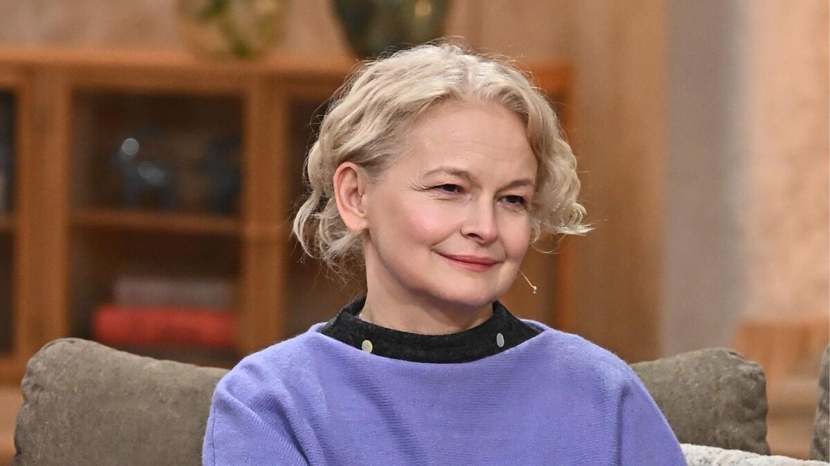 Dominika Ostałowska siedzi na kanapie w studiu programu „Pytanie na śniadanie” i z lekkim uśmiechem słucha rozmowy. Ma krótkie, jasne, lekko falowane włosy i fioletową bluzę zestawioną z ciemnym kołnierzem, a przy policzku widać mikroport. W tle znajduje się ciepło oświetlone, drewniane wnętrze studia z półkami i dekoracjami.