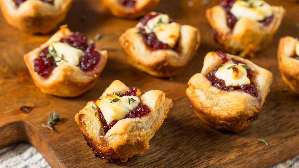 Kilka mini tart z ciasta francuskiego ustawionych na drewnianej desce. Każda porcja ma złociste, warstwowe ciasto wypełnione żurawiną i kawałkiem roztopionego oscypka. Wierzch posypany jest listkami tymianku, a tło pozostaje lekko rozmyte.