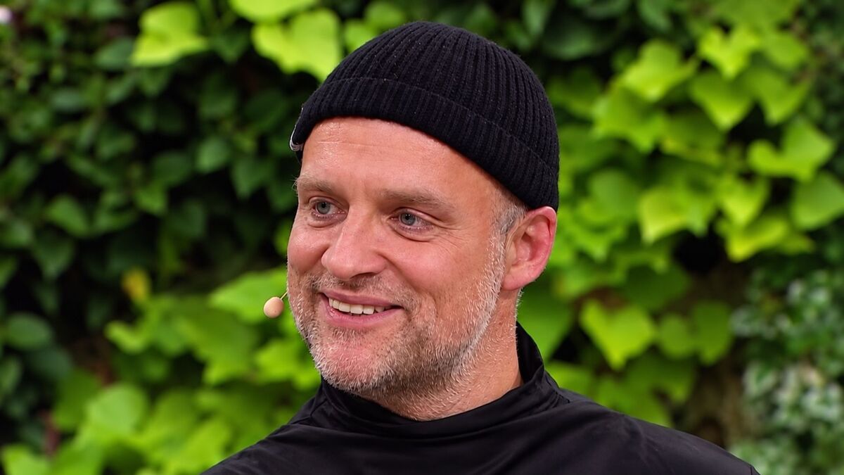Jarek Weber ma krótką brodę, czarną czapkę i spogląda lekko w bok. Jest ubrany w czarną bluzę z wysokim kołnierzem, a przy twarzy ma mikrofon nagłowny. W tle widoczne są gęste zielone liście.