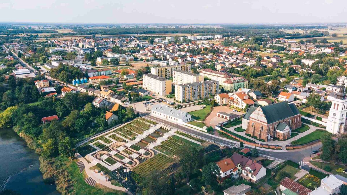 Uniejów został sfotografowany z lotu ptaka, pokazując zabudowę miasta oraz okoliczne tereny zielone. Na pierwszym planie widoczny jest kościół, park z geometrycznymi alejkami oraz fragment rzeki. W tle rozciągają się osiedla mieszkaniowe, pola i rozległa panorama okolicy.