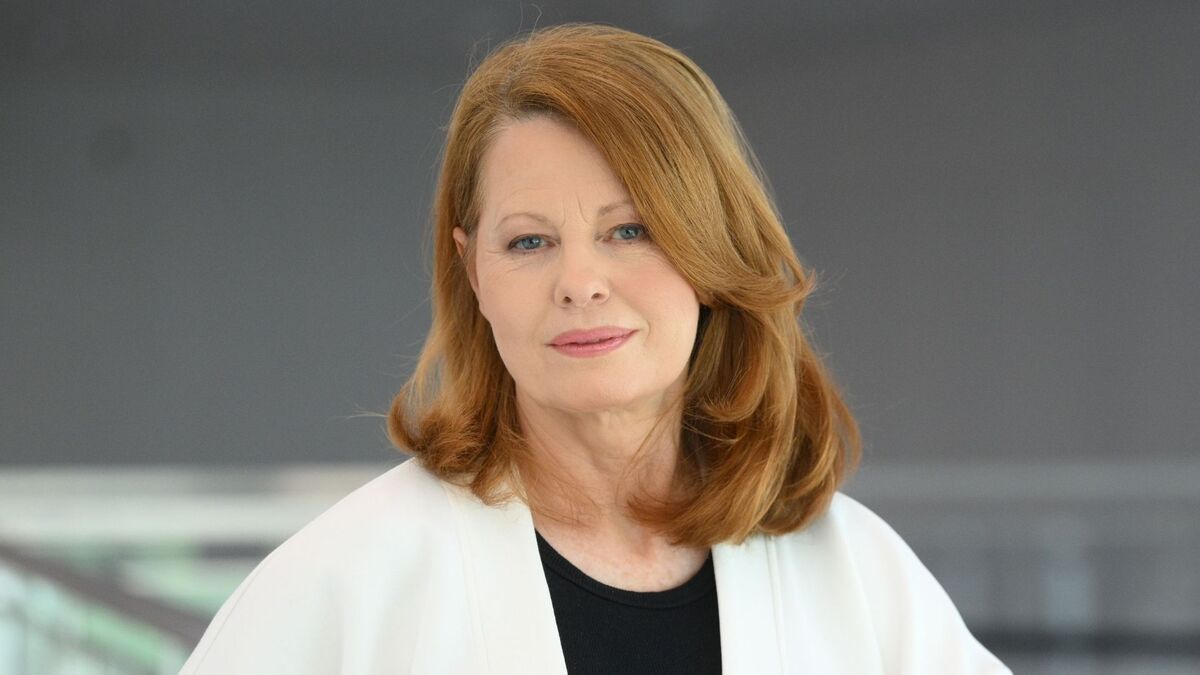 Ewa Skibińska pozuje do portretu w białej marynarce narzuconej na czarną bluzkę. Ma rude włosy sięgające ramion i patrzy wprost przed siebie z delikatnym uśmiechem. W tle znajduje się gładka szara powierzchnia oraz rozmyte jasne elementy wnętrza.