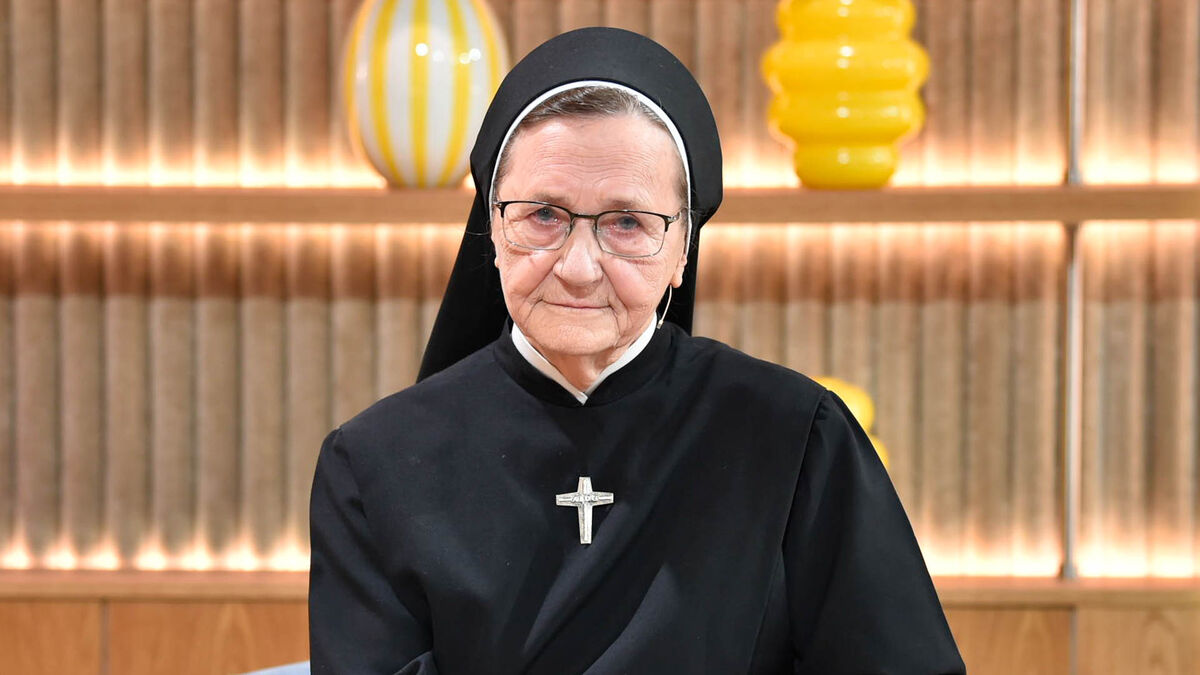 W studiu telewizyjnym widoczna jest starsza zakonnica w czarnym habicie i okularach, siedząca na fotelu. W tle znajdują się drewniane półki z dekoracyjnymi wazonami w jasnych kolorach.