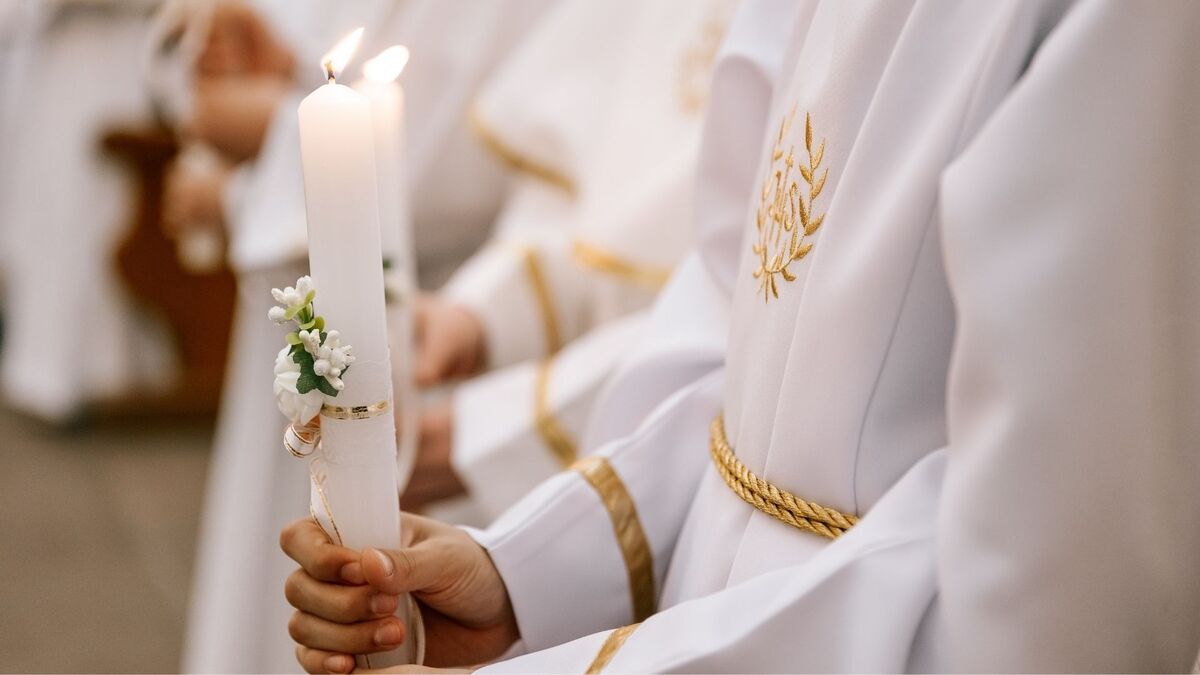 Zbliżenie na dziecko ubrane w białą albę z złotymi zdobieniami, trzymające zapaloną świecę komunijną. Świeca ozdobiona jest drobnymi białymi kwiatkami i złotą wstążką. W tle widać inne osoby w podobnych strojach, rozmyte poza głównym planem.